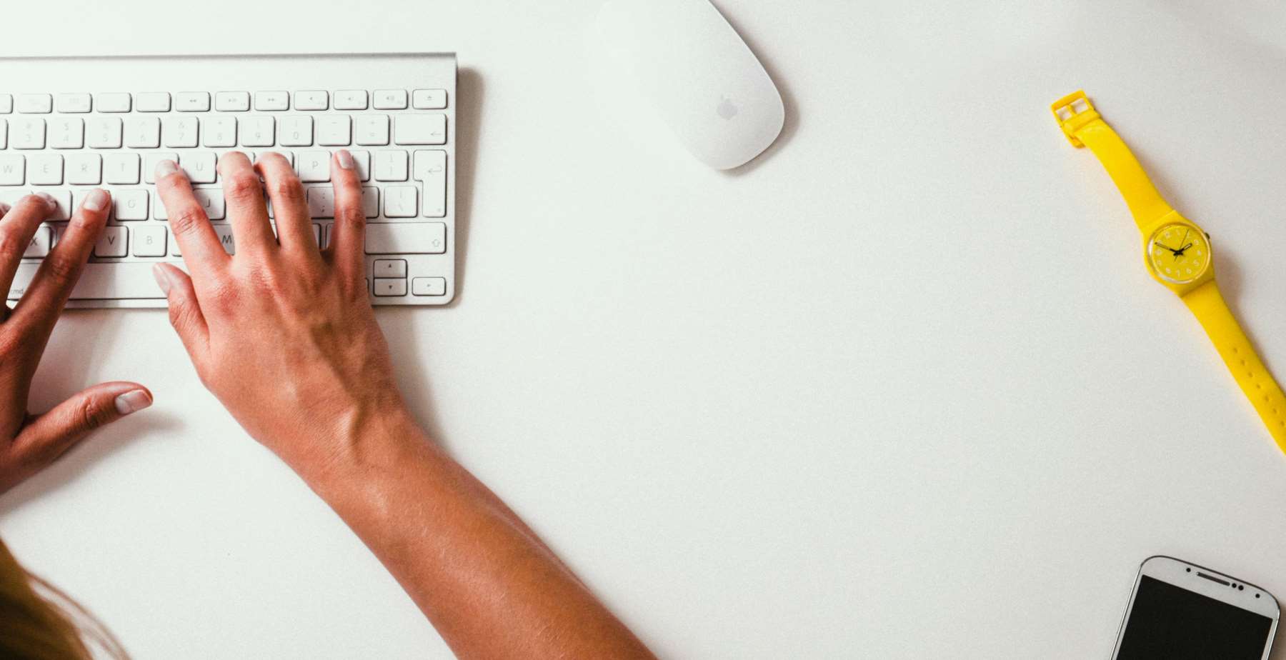 Person typing on a white keyboard beside a yellow watch and smartphone on a clean desk.